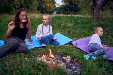 mother and her daughters  roasting and eating marshmallows on the campfire, camping vacation with children clipart