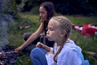 mother and her daughters  roasting and eating marshmallows on the campfire, camping vacation with children clipart