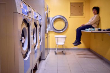      Genç ve cüsseli bir kadın, bir öğrenci yurdunun bodrumundaki çamaşır makinesine çamaşır yüklüyor.                          