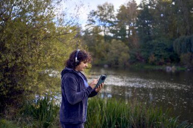 Orta yaşlı bir kadın, sonbahar parkında sabah koşusu sırasında telefonuyla kablosuz kulaklıkla müzik değiştiriyor.                        