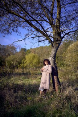 Orta yaşlı, siyah kıvırcık saçlı, açık bej takım elbiseli ve ceketli bir kadın sonbahar ormanında doğanın tadını çıkarıyor.                         