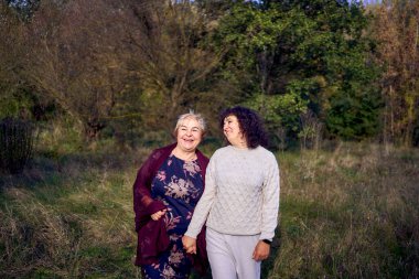 Orta yaşlı bir kız ve yaşlı bir anne birlikte sonbahar ormanında yürüyüş yapıyorlar.                   