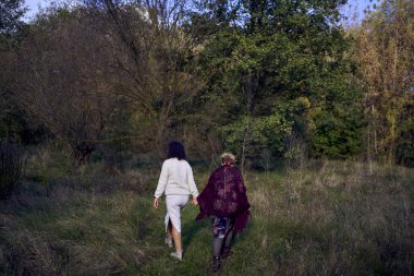 Orta yaşlı bir kız ve yaşlı bir anne birlikte sonbahar ormanında yürüyüş yapıyorlar.                   