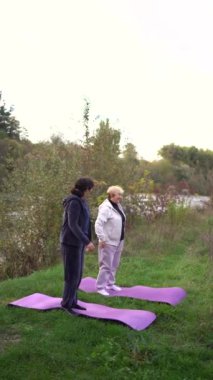 Orta yaşlı kadın, yaşlı annesine sonbaharda nehir kıyısında sağlığını korumak için somatik egzersizler ve yoga öğretiyor.