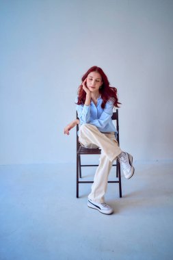 Beyaz arka planda bir sandalyede oturan kızıl saçlı güzel bir genç kızın portresi.                     