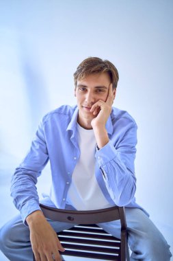 Mavi gömlekli, mavi arka planda bir sandalyede oturan genç ve yakışıklı bir adamın portresi.                           