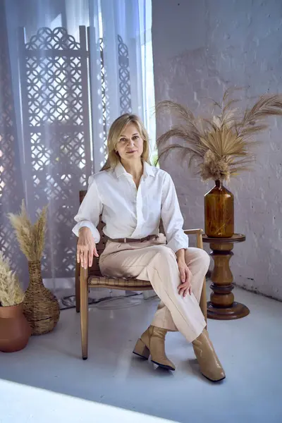 Beyaz bluz ve bej pantolon giymiş orta yaşlı şık bir kadının portresi.                    