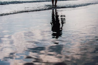 Adam kumsalda yürüyor, ıslak kumda yansıyor.