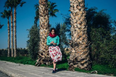Parlak elbiseli genç bir kadın palmiye ağaçlarının önünde duruyor.