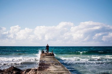 Rıhtımdaki genç bir adamın portresi, dalgalar sıçrıyor ve iskeleye çarpıyor.