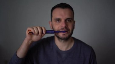 30 yaşındaki bir adam elektrikli diş fırçası kullanarak dişlerini mor diş macunuyla fırçalıyor.