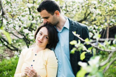 Bir koca ve hamile bir eş, çiçek açan elma ağaçlarıyla çevrili bir bahar bahçesinde sarılıyorlar.