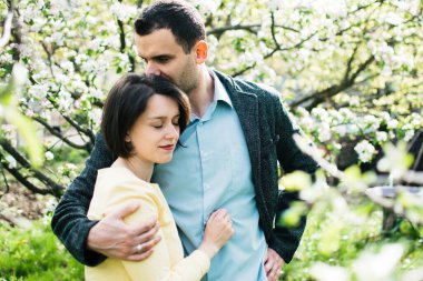 Bir koca ve hamile bir eş, çiçek açan elma ağaçlarıyla çevrili bir bahar bahçesinde sarılıyorlar.