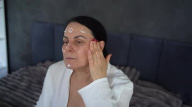 Yatakta iç çamaşırlı yaşlı bir kadın yüz ve vücut estetiğiyle rutin bir kişisel bakım sergiliyor.