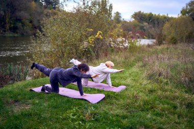  Orta yaşlı kadın, yaşlı annesine sonbaharda nehir kıyısında sağlığını korumak için somatik egzersizler ve yoga öğretiyor.                         