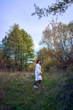 Orta yaşlı, siyah kıvırcık saçlı, açık bej takım elbiseli bir kadın sonbahar ormanında doğanın tadını çıkarıyor.                