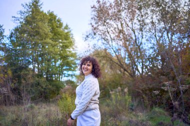 Orta yaşlı, siyah kıvırcık saçlı, açık bej takım elbiseli bir kadın sonbahar ormanında doğanın tadını çıkarıyor.                