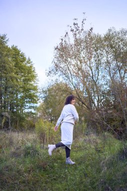Orta yaşlı, siyah kıvırcık saçlı, açık bej takım elbiseli bir kadın sonbahar ormanında doğanın tadını çıkarıyor.                