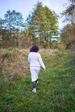 Orta yaşlı, siyah kıvırcık saçlı, açık bej takım elbiseli bir kadın sonbahar ormanında doğanın tadını çıkarıyor.                