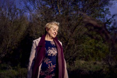          Gri saçlı, açık bej ceketli yaşlı kadın sonbahar ormanlarında doğanın tadını çıkarıyor.                      