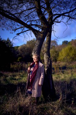          Gri saçlı, açık bej ceketli yaşlı kadın sonbahar ormanlarında doğanın tadını çıkarıyor.                      