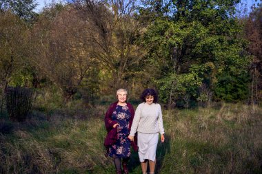 Orta yaşlı bir kız ve yaşlı bir anne birlikte sonbahar ormanında yürüyüş yapıyorlar.                   