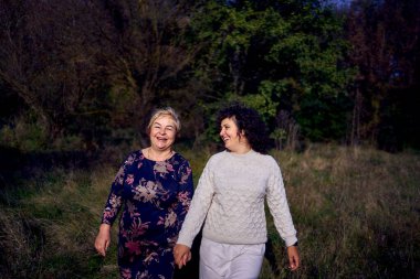 Orta yaşlı bir kız ve yaşlı bir anne birlikte sonbahar ormanında yürüyüş yapıyorlar.                   