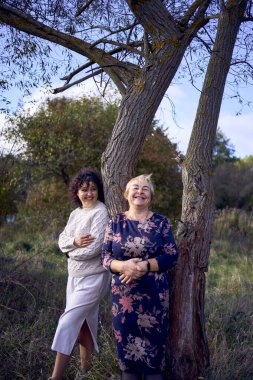 Orta yaşlı bir kız ve yaşlı bir anne birlikte sonbahar ormanında yürüyüş yapıyorlar.                   