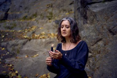 a   woman in a black long dress sits on black rocks on Halloween holding a burning candle, performs a witch ritual, modern witch                         