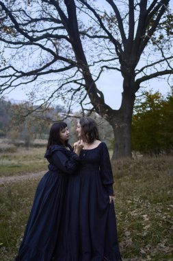      Two women in long black dresses perform a historical reenactment of the Edwardian era dancing lovers on a hill near a majestic oak tree for Halloween.                          