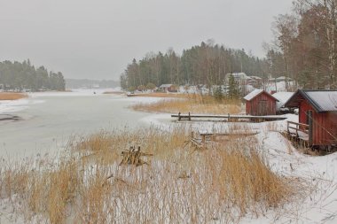 Deniz boğazının kış manzarası, deniz kenarındaki kulübeler, sazlıklar, ağaçlar, bulutlu kış havasında deniz buzu ve su, Pellinki, Finlandiya.