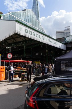 Londra, İngiltere 'de güneşli bir günde arka planda Parça' nın olduğu Borough Market 'in girişi. Kabinler önde görünüyor ve insanlar arka planda alışveriş yapıyor..