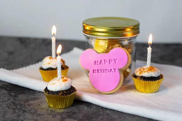 Happy Birthday pink mouse cookie propped up on a gold coloured jar with a chocolate cupcake with white marshmallow icing, edible gold leaf on the top and gold wrapper - 45 degree angle shot