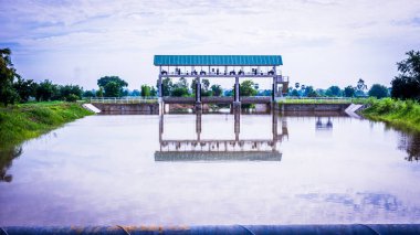 Görkemli bir mavi su kapısı berrak bir mavi gökyüzüne karşı dimdik ayakta duruyor, yansıması durgun suda yansıtılıyor, sakin ve resimsel bir manzara yaratıyor. Su kapıları, sellerin önlenmesi, yaşamların ve mülklerin korunmasında hayati rol oynar..