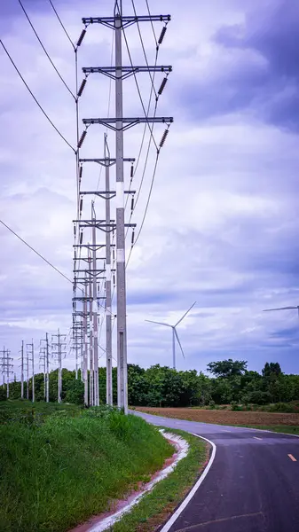 stock image High-voltage electric poles transmit power along the roadside, integrated with a wind turbine clean energy project, showcasing the synergy between traditional and renewable energy sources.																									