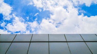 Doğa ve mimarinin sakin bir birlikteliği: Binanın gri cephesi açık mavi gökyüzü ve dalgalanan bulutlarla keskin bir tezat oluşturuyor.