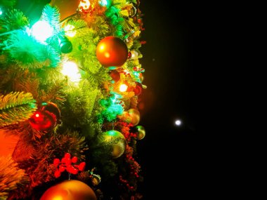 Karanlık bir gece göğüne karşı parıldayan renkli ışıklar ve süslemelerle süslenmiş canlı bir Noel ağacı. Bayram süslemeleri arasında kırmızı, altın ve yeşil mücevherler de yer alıyor ve sıcak bir tatil ortamı yaratıyor..