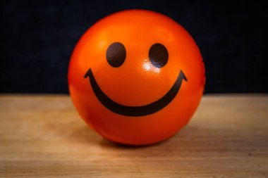 Parlak turuncu top, gülen yüzlü, ahşap zemin üzerine koyu arka plana yerleştirilmiş. Neşeli ve canlandırıcı bir görsel ruh hali veya duygu tasarımı için mükemmel..