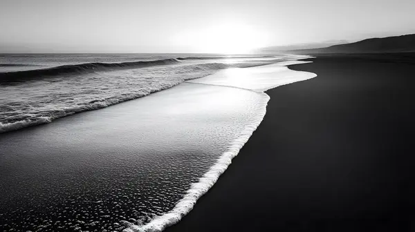 Şafak vakti terk edilmiş bir plajın siyah-beyaz görüntüsü, yumuşak dalgalar ve ışığın etkileşimi ve gölgenin barışçıl, zamansız bir sahne yaratması.