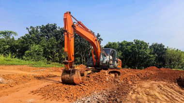 Bir yol inşaatında çalışan turuncu bir kazıcı. Bol miktarda kırmızı toprak ve arka planda açık mavi gökyüzü olan ezilmiş taşlar.