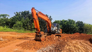 Bir yol inşaatında çalışan turuncu bir kazıcı. Bol miktarda kırmızı toprak ve arka planda açık mavi gökyüzü olan ezilmiş taşlar.