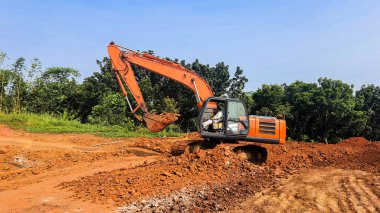 Bir yol inşaatında çalışan turuncu bir kazıcı. Bol miktarda kırmızı toprak ve arka planda açık mavi gökyüzü olan ezilmiş taşlar.