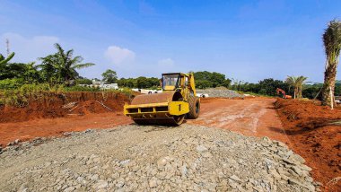 Yol yapımı projesinde çalışan sarı tekerlekli bir inşaat makinesi. Bol miktarda kırmızı toprak ve arka planda açık mavi gökyüzü olan ezilmiş taşlar.