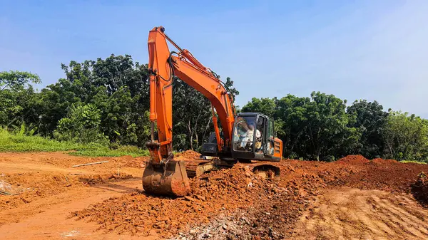 Bir yol inşaatında çalışan turuncu bir kazıcı. Bol miktarda kırmızı toprak ve arka planda açık mavi gökyüzü olan ezilmiş taşlar.