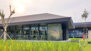 Jakarta Nov 01 2023: Cam ve desenli seramik cepheli ve önünde uzun çimenler olan bir bina. Pirinç tarlasına benziyor. Güzel peyzaj mimarisi. Günbatımı gökyüzü, insan yok. Yansıyan gölet