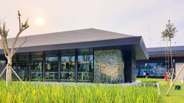 Jakarta Nov 01 2023: Cam ve desenli seramik cepheli ve önünde uzun çimenler olan bir bina. Pirinç tarlasına benziyor. Güzel peyzaj mimarisi. Günbatımı gökyüzü, insan yok. Yansıyan gölet