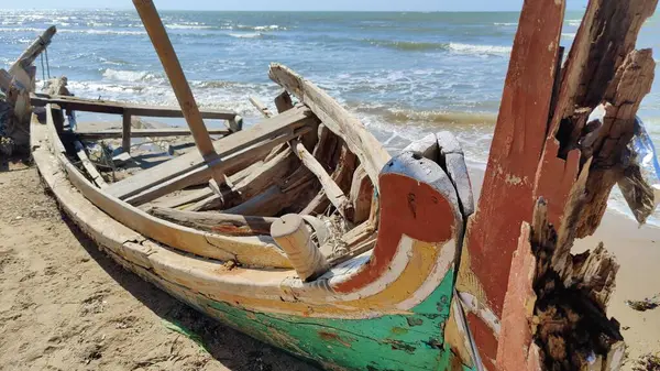 A broken fishing boat, with its hull partially stranded on the edge of a white sand beach.