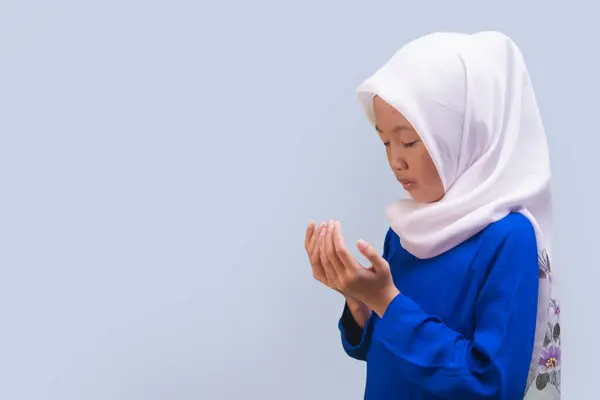 Side view of Little muslim girl in traditional clothes praying Dua with duck Egg Blue on background