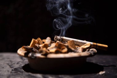 Sigaralar kül tablasında dumanla yanıyor, siyah arka planda.