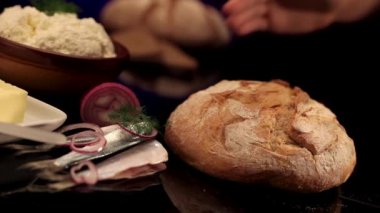 Bir kadının kabuklu ekmeği alıp tekrar siyah cam masaya koyduğu statik pozlar. Etraftaki diğer tabaklar, balıklar, soğanlar, tereyağı, süzme peynir.